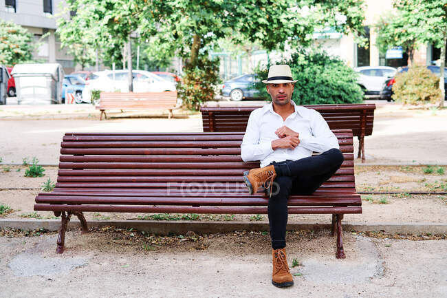 Hombre hispano joven guapo y seguro en ropa elegante y sombrero sentado en el banco y mirando hacia otro lado mientras descansa en la calle de la ciudad - foto de stock