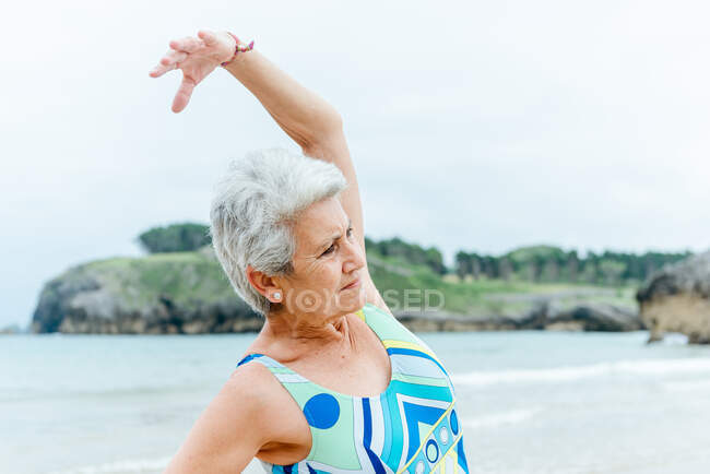 Feminino sênior positivo em maiô elegante fazendo exercício de curvatura lateral enquanto pratica um estilo de vida saudável e exercita-se na praia contra o mar ondulado — Fotografia de Stock
