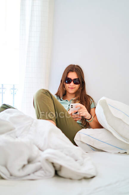 Smiling young female with sunglasses sitting on bed and browsing on smartphone while spending time alone at home — Stock Photo