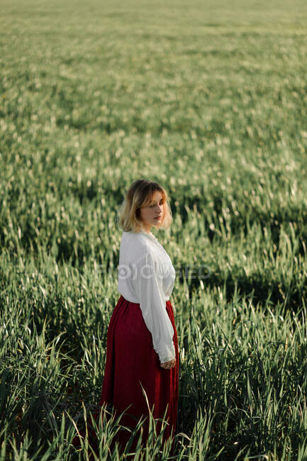 Calma joven hembra vestida con blusa y falda anticuadas de pie solo entre la hierba verde alta en el día nublado de verano en el campo - foto de stock