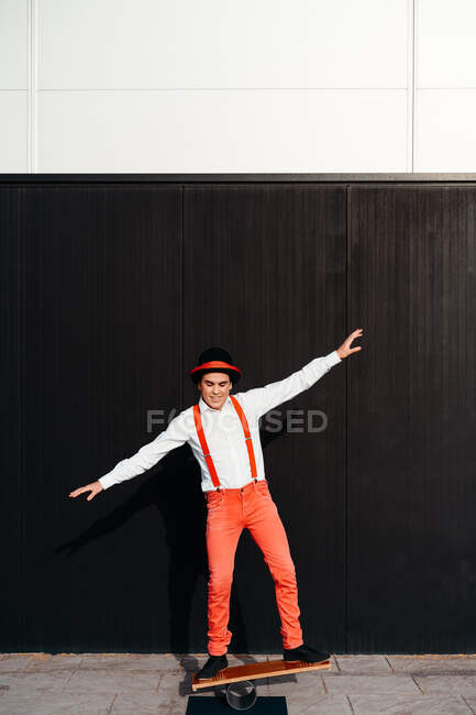 Corpo inteiro de artista de circo masculino em calças vermelhas e chapéu realizando truque no tabuleiro de equilíbrio contra a parede preta — Fotografia de Stock