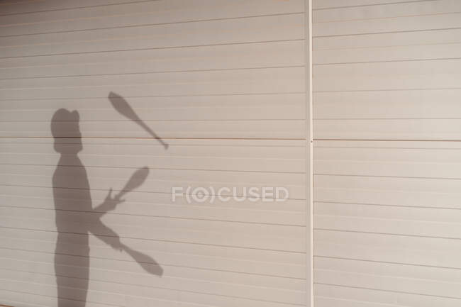 Side view shadow of anonymous talented male circus performer in hat juggling clubs near white wall — Stock Photo