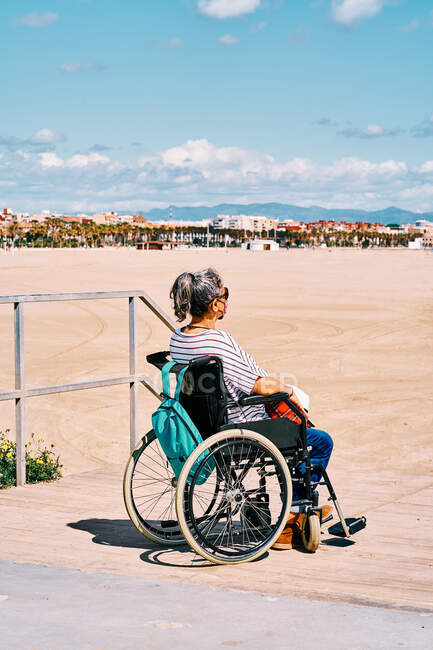 Вид сбоку женщины в защитной маске, сидящей в инвалидной коляске с рюкзаком и наслаждающейся солнечным летним днем на песчаном пляже — стоковое фото