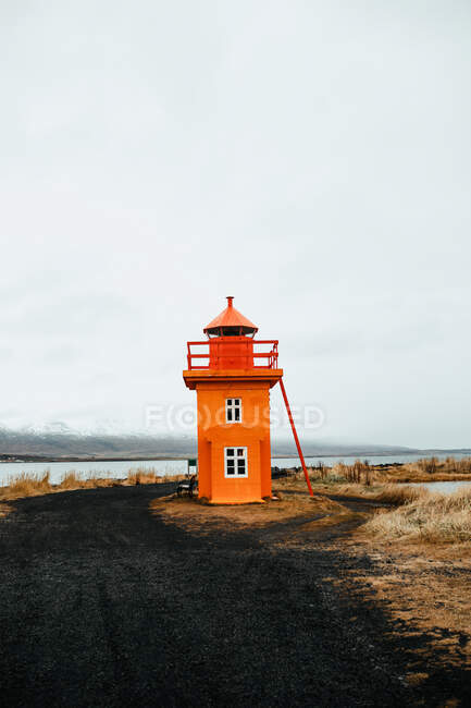Orange lighthouse on coast between wild lands near water surface and cloudy sky — Stock Photo