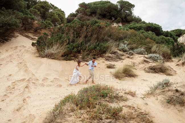 Delighted multiracial bridge and groom holding hands and running along sandy hill on wedding day in nature — Stock Photo