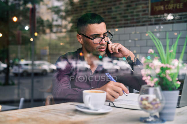 Kluger hispanischer Geschäftsmann mit Brille, der Notizen im Notizblock macht und Anrufe entgegennimmt, während er hinter dem Fenster in der Cafeteria sitzt — Stockfoto