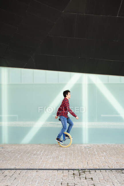 Visão lateral de corpo inteiro do jovem homem ativo em camisa quadriculada e jeans executando truque em monociclo perto de parede de vidro espelhado do edifício contemporâneo — Fotografia de Stock