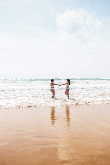 Side view of cheerful female friends holding hands in swimsuits in foamy ocean near sandy beach under blue cloudy sky in sunny day — Stock Photo