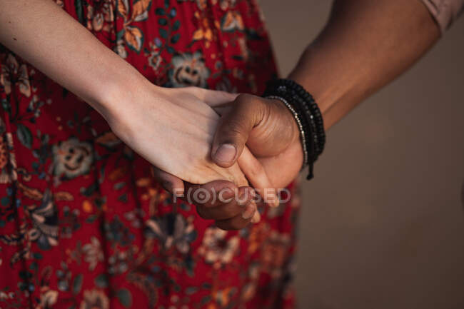 Анонимная женщина в красочном платье и черный мужчина с браслетом на запястье, держась за руки, наслаждаясь романтическими моментами вместе — стоковое фото