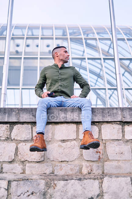 From below full body of modern young male in trendy casual outfit with smartphone in hand looking away while sitting on stone fence against urban building — Stock Photo