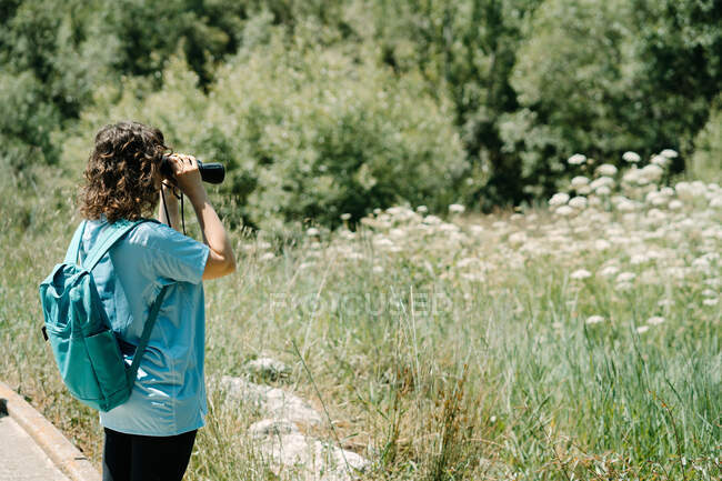 Вид збоку на невпізнавану жінку-добровольцю в блакитній сорочці з рюкзаком, що досліджує природу через бінокль під час екологічної кампанії в літньому лісі — стокове фото