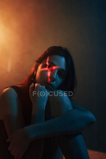 Jovem modelo feminino sem emoção com projeção de luz em forma de cruz no rosto sentado no estúdio escuro e olhando para a câmera — Fotografia de Stock