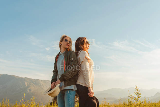 Junge enge Freundinnen in schicken Klamotten stehen zusammen auf einer Wiese in den Bergen und schauen im goldenen Licht weg — Stockfoto