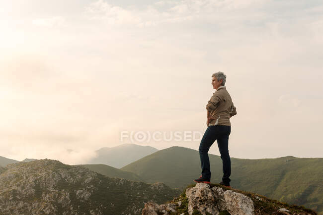 Vista lateral da exploradora idosa empoderada com as mãos na cintura admirando o terreno montanhoso contra o céu nublado do nascer do sol de manhã na natureza — Fotografia de Stock