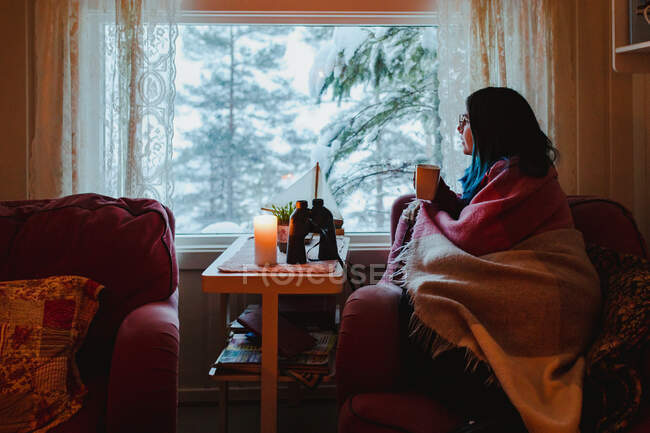 Бічний вид молодої жінки в окулярах під ковдрою відпочиває на дивані з чашкою біля вікна з видом на яловичину лісу в снігу. — стокове фото