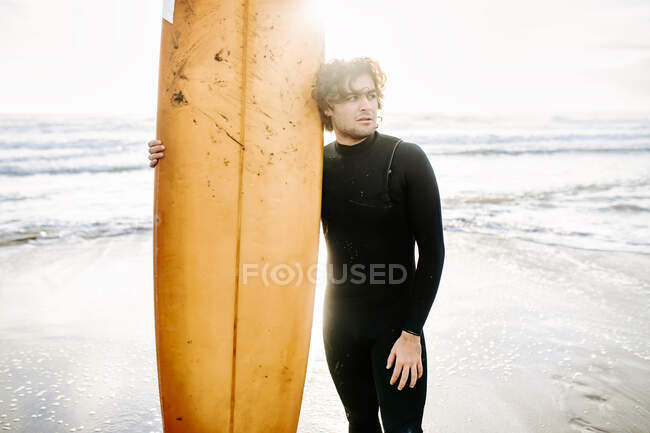 Surfer uomo vestito in muta in piedi guardando lontano con la tavola da surf sulla spiaggia durante l'alba sullo sfondo — Foto stock