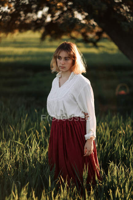 Спокійна молода жінка, одягнена в старомодну блузку і спідницю, що стоїть один серед високої зеленої трави в похмурий літній день у сільській місцевості — стокове фото