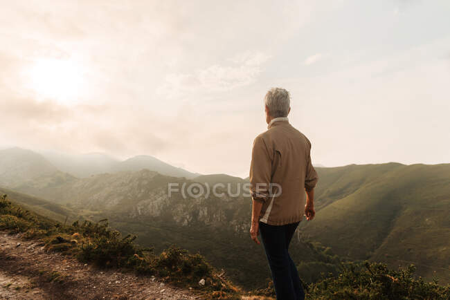 Задний вид стоящего анонимного исследователя, любующегося горной местностью против облачного восхода солнца утром в природе — стоковое фото