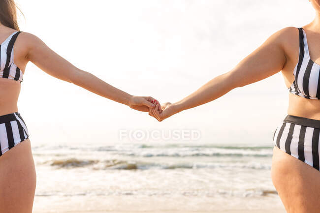 Безликие девушки в купальниках, держащиеся за руки на песчаном побережье возле волнистого моря в солнечный день под облачным небом — стоковое фото