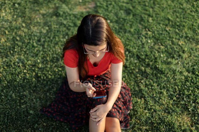 Donna in abiti estivi seduta sul prato verde nel parco e navigare in Internet sul cellulare mentre intrattenere durante il fine settimana di sera — Foto stock