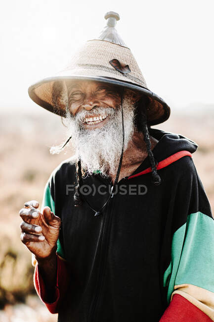 Fröhlicher alter Rastafari mit Dreadlocks, der in die Kamera schaut, während er auf einer trockenen Wiese in der Natur Unkraut raucht — Stockfoto
