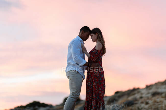 Liebendes Paar in eleganter Kleidung, das sich auf einem Hügel vor dem Hintergrund des Sonnenuntergangs über dem Meer im Sommer umarmt — Stockfoto