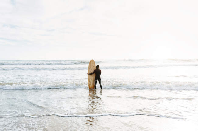 Обратный вид на неузнаваемого серфера, одетого в гидрокостюм, стоящего, смотрящего с доской для серфинга в сторону воды, чтобы поймать волну на пляже во время восхода солнца — стоковое фото