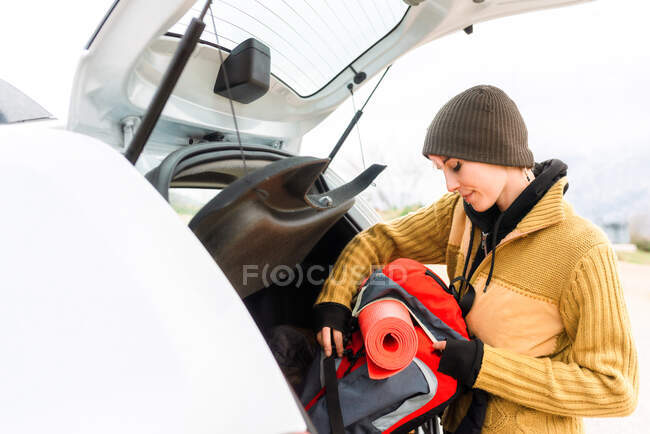 Vista laterale del contenuto avventuriero donna in piedi con zaino da viaggio vicino al bagagliaio dell'auto in montagna — Foto stock