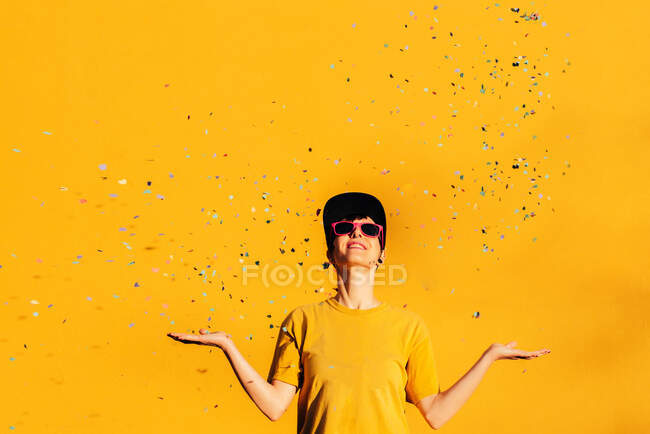 Современная женщина в хип-хоп кепке и солнцезащитных очках раздувает красочные конфетти и веселится на жёлтом фоне — стоковое фото