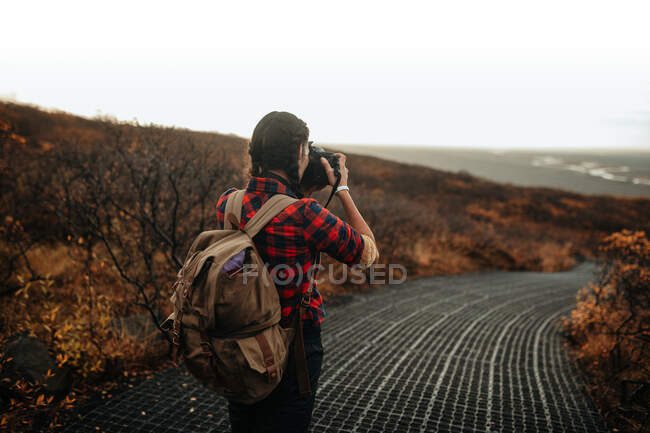 Vista trasera del joven turista en la pasarela tomando fotos del valle desde la colina y el cielo nublado - foto de stock