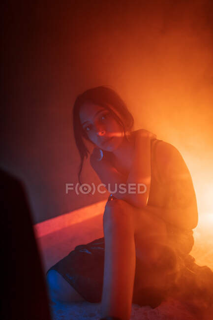 Tranquil young female model in dress sitting on floor and leaning on hand while looking at camera in dark studio with colorful lights — Stock Photo