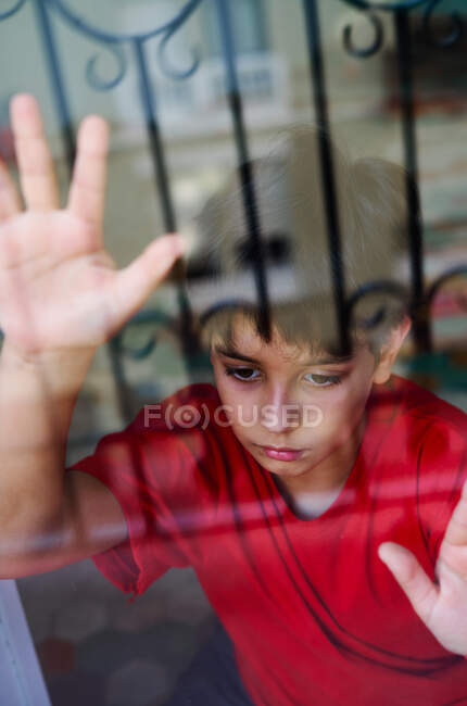 Through glass of unhappy preteen boy with bruises on face looking away while standing near window at home as concept of domestic violence and child abuse — Stock Photo