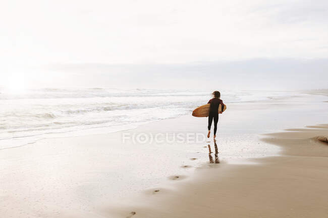 Обратный вид на неузнаваемого серфера в гидрокостюме, бегущего с доской для серфинга к воде, чтобы поймать волну на пляже во время восхода солнца — стоковое фото