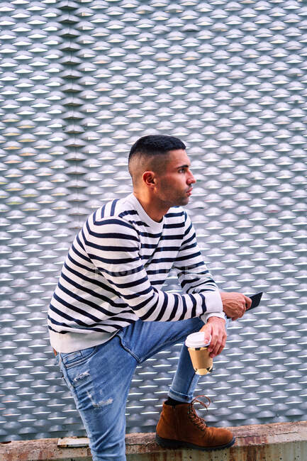 Homem hispânico em roupa elegante olhando para longe e usando o celular enquanto se inclina na parede na rua da cidade — Fotografia de Stock