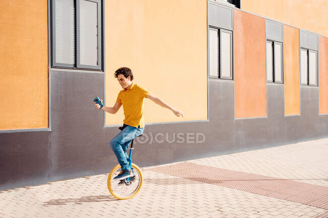 Corps complet de jeune homme agile utilisant le téléphone mobile tout en chevauchant monocycle près de bâtiment urbain coloré moderne — Photo de stock