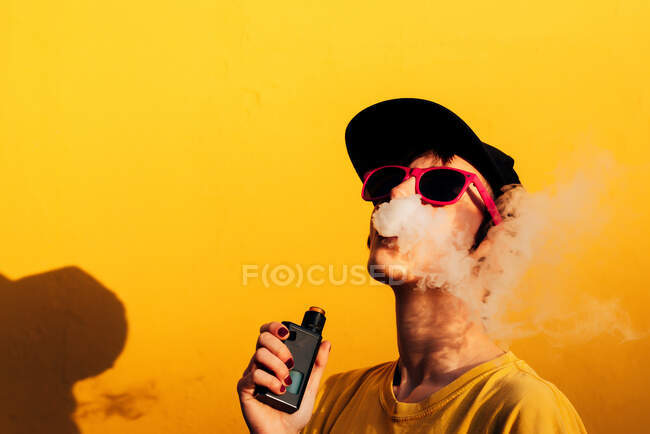 Femme contemporaine dans une tenue élégante expirer les fumées tout en se tenant près du mur jaune et vaping sur la rue de la ville — Photo de stock