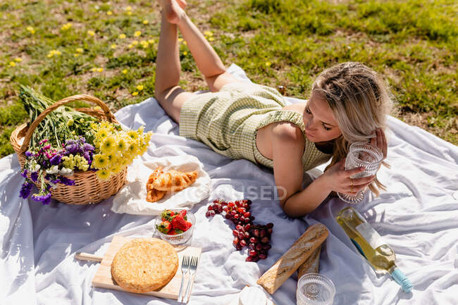 Alto ángulo de hembra acostada sobre manta con copa de vidrio cerca de fresas en tazón y uvas cerca de botella de vino colocada cerca de baguette y focaccia - foto de stock