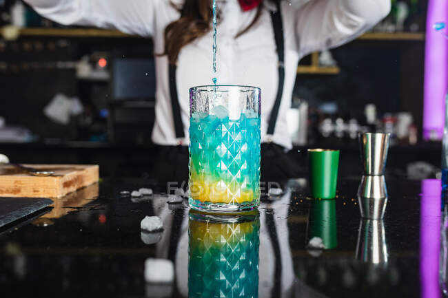 Beschnitten bis zur Unkenntlichkeit weibliche Barkeeper in stilvollem Outfit serviert blauen Cocktail aus einem Shaker in ein Glas, während an der Theke in modernen Bar stehen — Stockfoto