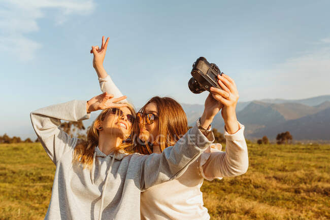 Joyeux jeunes copines dans les lunettes de soleil prendre soi-même photo avec caméra de film analogique montrant des signes V dans la campagne des montagnes — Photo de stock
