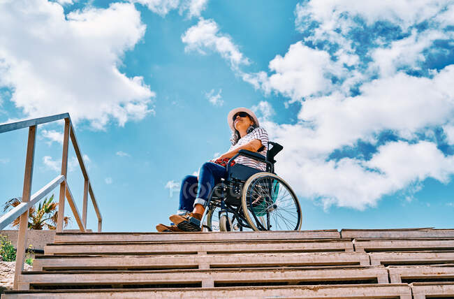 Bajo ángulo cuerpo completo de hembra discapacitada positiva sentada en silla de ruedas cerca de la escalera mirando hacia otro lado contra el cielo azul en la ciudad - foto de stock