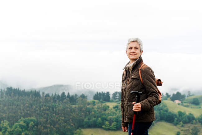 Smiling elderly woman with backpack and trekking stick standing on grassy slope towards mountain peak during trip in nature looking away — Stock Photo