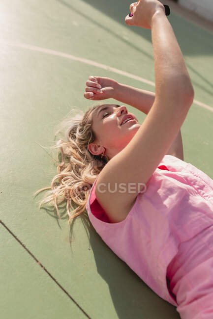 Young glad female in pink dress taking selfie on instant photo camera while lying on ground on sunny day — Stock Photo