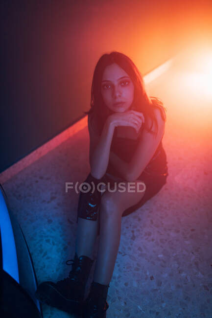 From above of tranquil young female model in dress sitting on floor and leaning on hand while looking at camera in dark studio with colorful lights — Stock Photo