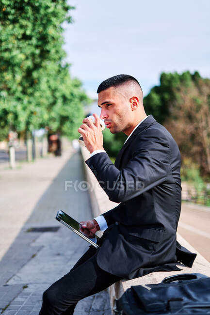 Low angle side view of young Hispanic male in formal black suit with mobile phone in hand enjoying refreshing takeaway drink while resting on urban street in summer day — Stock Photo