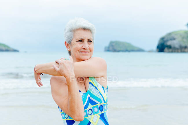 Положительный седой пенсионерка в купальнике растягивая плечи во время фитнес-тренировки на пляже возле размахивая морем в летний день — стоковое фото