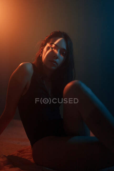 Молода неемоційна жіноча модель з хрестоподібною світловою проекцією на обличчі, сидячи в темній студії і дивлячись на камеру — стокове фото