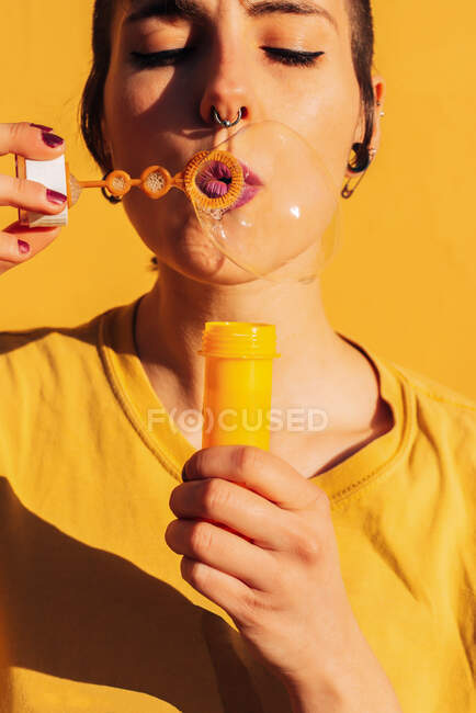Cortar fêmea moderna com piercing soprando bolhas de sabão com os olhos fechados na câmera no dia ensolarado contra a parede amarela — Fotografia de Stock