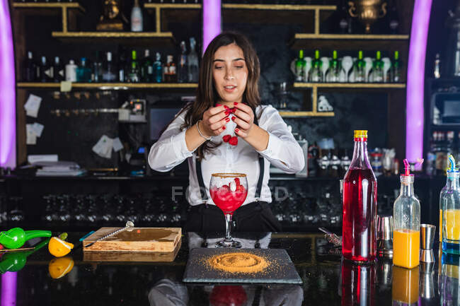 Молода жінка-банкір у стильному вбранні, що прикрашає коктейль з малиною, стоячи за стійкою в сучасному барі — стокове фото