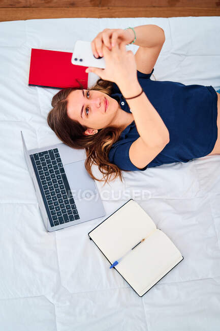 Зверху молода студентка лежить на ліжку з ноутбуком і підручниками, беручи автопортрет на смартфон під час дистанційних онлайн-навчань вдома — стокове фото