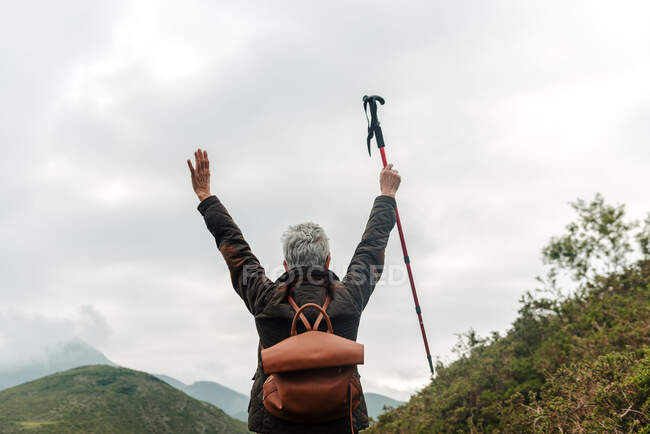 Visão traseira da mulher idosa anônima com mochila segurando a bengala nos braços levantados contra o céu cinza nublado enquanto explora a natureza — Fotografia de Stock